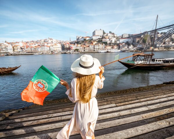 Young woman traveler standing back with portuguese flag, enjoying beautiful cityscape view on Douro river and boats during the morning light in Porto, Portugal