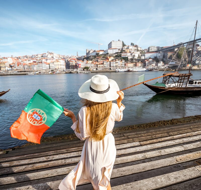 Young woman traveler standing back with portuguese flag, enjoying beautiful cityscape view on Douro river and boats during the morning light in Porto, Portugal
